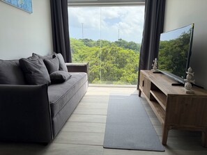 A modern living room with a grey sofa, a wooden TV stand, and a view of greenery through a large window.