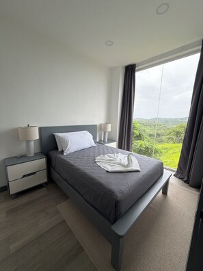 Un dormitorio moderno con una cama grande, dos mesitas de noche y una mesita auxiliar, con vistas a un paisaje exuberante.