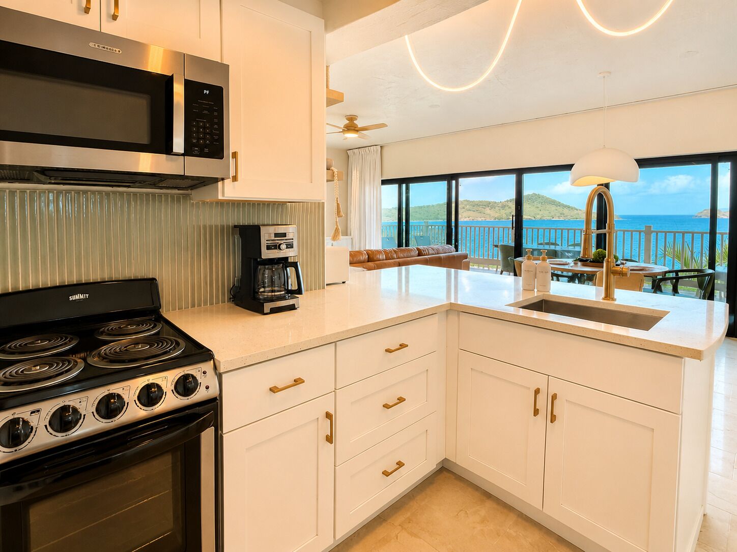 Una cocina moderna con estufa integrada, microondas y cafetera. Cuenta con armarios blancos con detalles dorados y un amplio ventanal con vistas panorámicas.