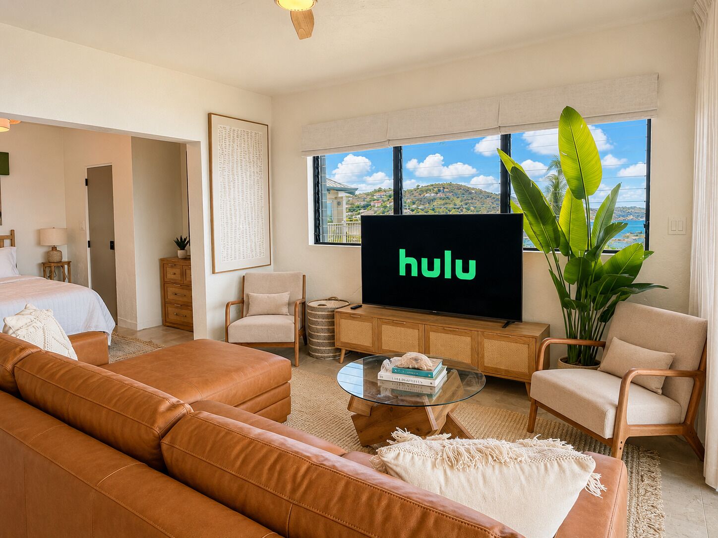 Een moderne hotelkamer met een groot bed, een bruine leren bank, een glazen salontafel, een televisiescherm met het Hulu logo en uitzicht op zee.