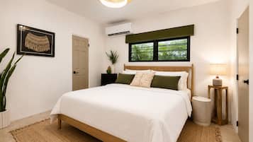 A bedroom with a bed, a bedside lamp, a potted plant, and a window with a view of greenery.