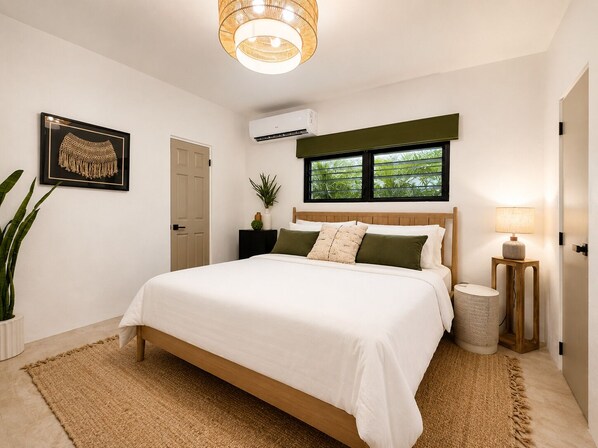 A bedroom with a bed, a bedside lamp, a potted plant, and a window with a view of greenery.