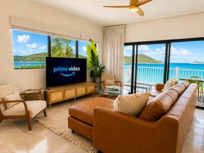 A living room with a large flat-screen TV, a leather sofa, a coffee table, and a view of the ocean.