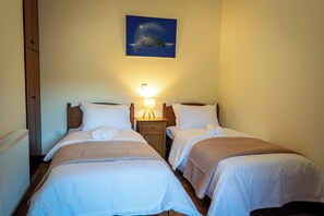 Quarto de hotel com duas camas, mesa de cabeceira com abajur, quadro na parede e radiador.