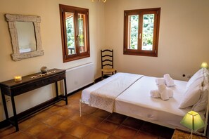 Ein Schlafzimmer mit einem Bett, einem Nachttisch mit Lampe, einem Sessel und zwei Fenstern mit Blick ins Grüne.