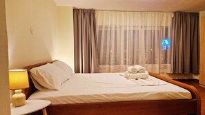 Een hotelkamer met een bed, een bedlampje, een raam met gordijnen en uitzicht op de stad 's nachts.