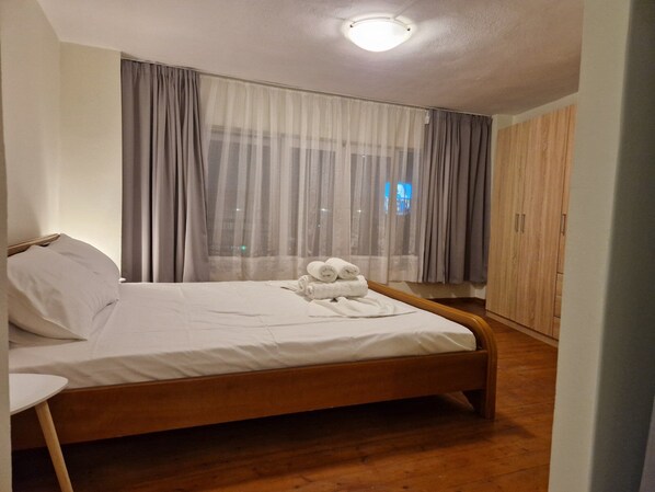 Een slaapkamer met een houten bed, witte lakens, een groot raam met dunne gordijnen en een kledingkast.