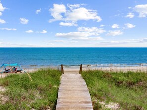 Une promenade en bois qui mène à une plage, avec un ciel bleu dégagé et quelques nuages.
