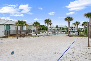 Un terrain de beach-volley avec un filet, des palmiers et des maisons de plage en arrière-plan.