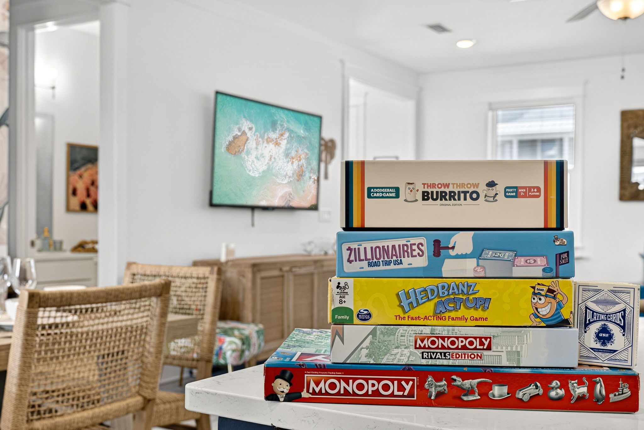 A stack of board games on a table in a room with a dining area and a painting on the wall.