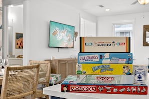 A stack of board games on a table in a room with a dining area and a painting on the wall. - New! 5BR Beach Family Retreat, Pool, Water Views, Pickleball, Dog-Friendly (Port St. Joe)