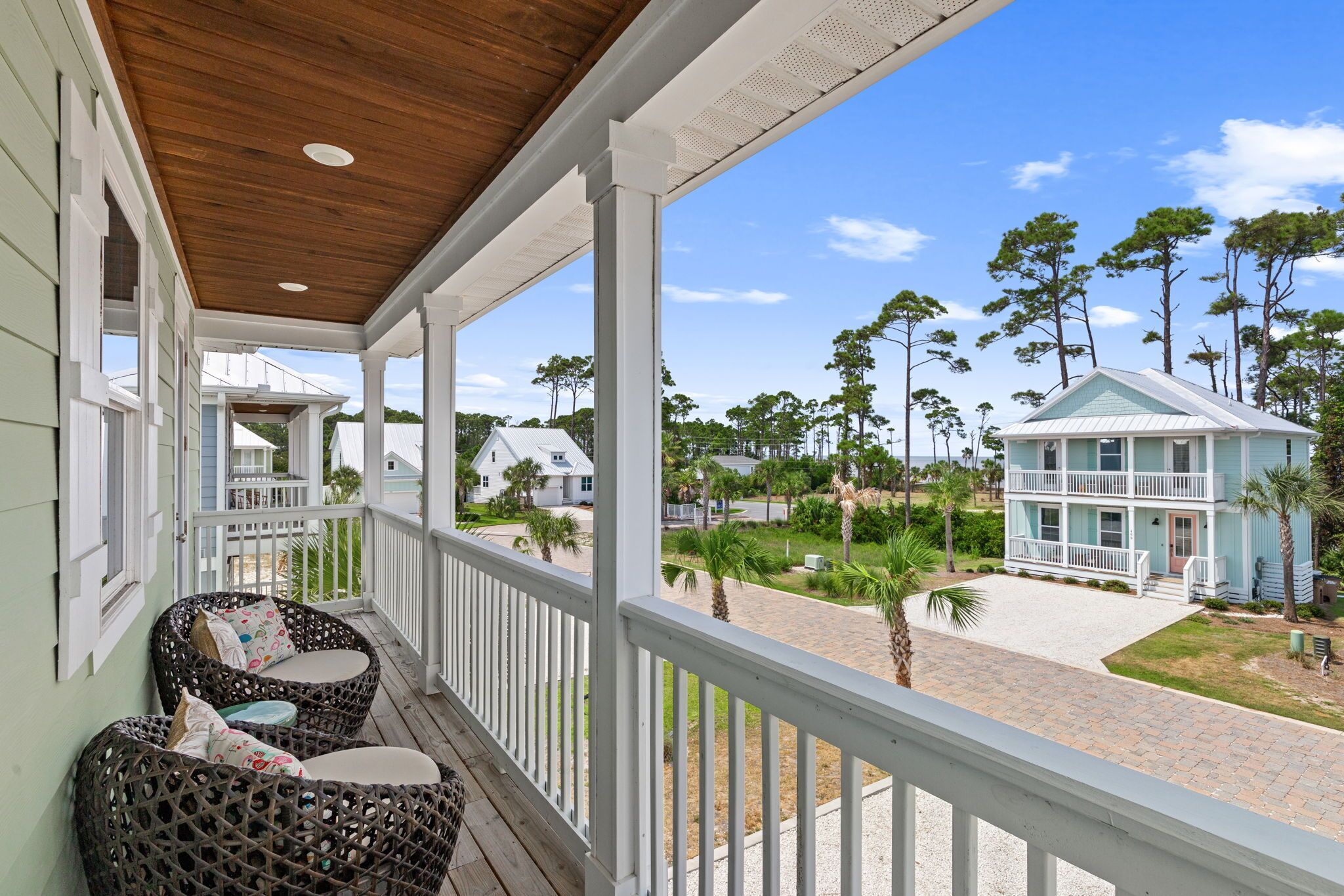 A covered porch with wicker furniture, a view of a residential area with houses and trees, and a clear sky.