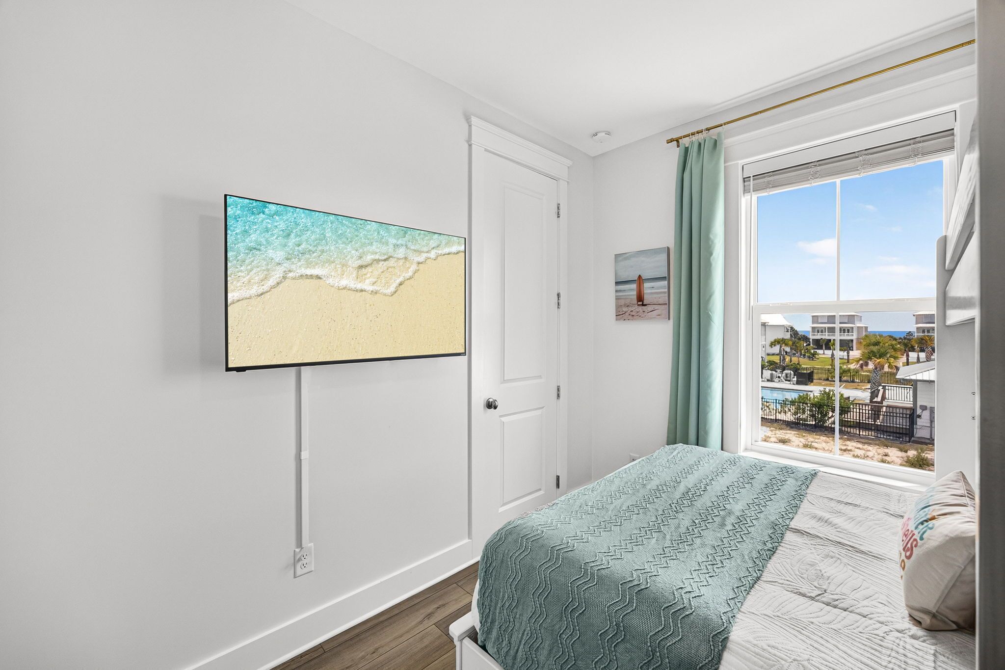 A bedroom with a bed, a wall-mounted TV, a door, and a window with a view of a building and a beach.
