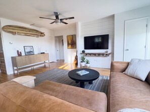 A living room with a sofa, a coffee table, a TV, and a ceiling fan. - Welcome to Golden Hour Bungalow — a cozy, mid-century surf retreat. (Carolina Beach)