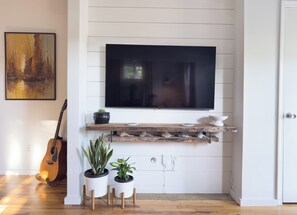 A room with a mounted TV, wooden shelves, potted plants, and a guitar. - Welcome to Golden Hour Bungalow — a cozy, mid-century surf retreat. (Carolina Beach)