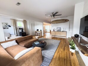 A modern living room with a sofa, coffee table, and a TV. There is a wooden floor and a ceiling fan. - Welcome to Golden Hour Bungalow — a cozy, mid-century surf retreat. (Carolina Beach)