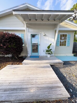 A house with a wooden deck leading to a front door with a blue door and matching shutters, a small porch with a light fixture, and a potted plant beside the door. - Welcome to Golden Hour Bungalow — a cozy, mid-century surf retreat. (Carolina Beach)