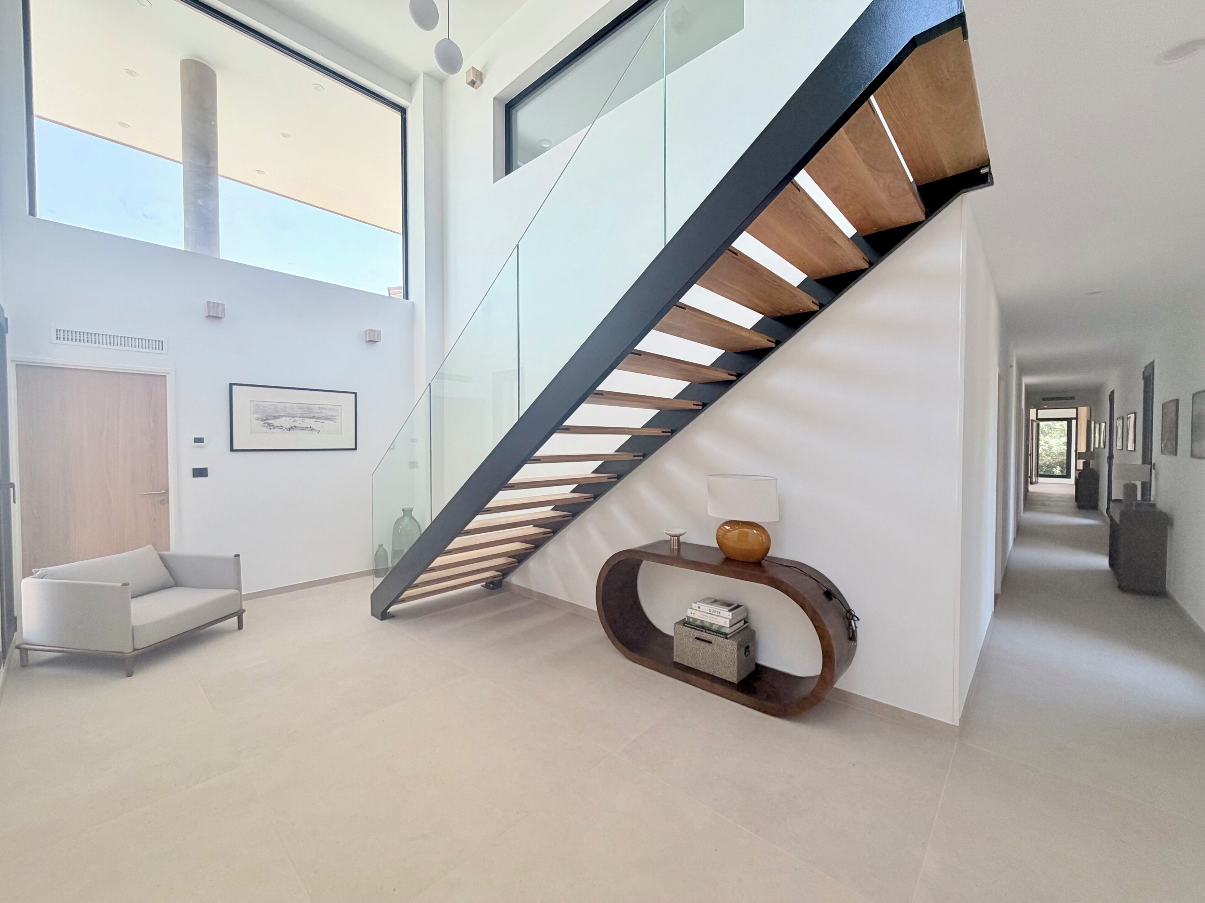 A modern interior with a glass staircase, a white sofa, and a hallway with framed pictures.