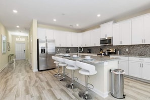 A modern kitchen with white cabinets, a stainless steel refrigerator, a granite countertop, and a bar area with stools. - Spacious 6-bedroom 5 bathroom house in Champions Gate With Pool & Spa (Four Corners)