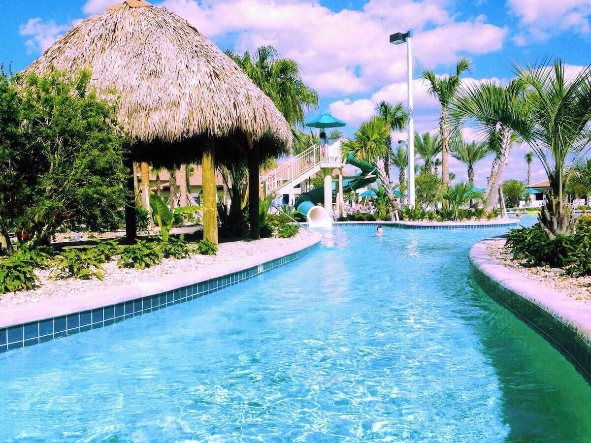 A swimming pool with a thatched-roof structure, palm trees, and a clear blue sky.