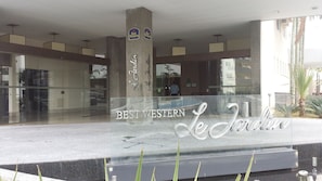 Entrada de hotel com fachada de vidro, placa com a inscrição 'BEST WESTERN Le Jardin' e uma pessoa visível no interior.