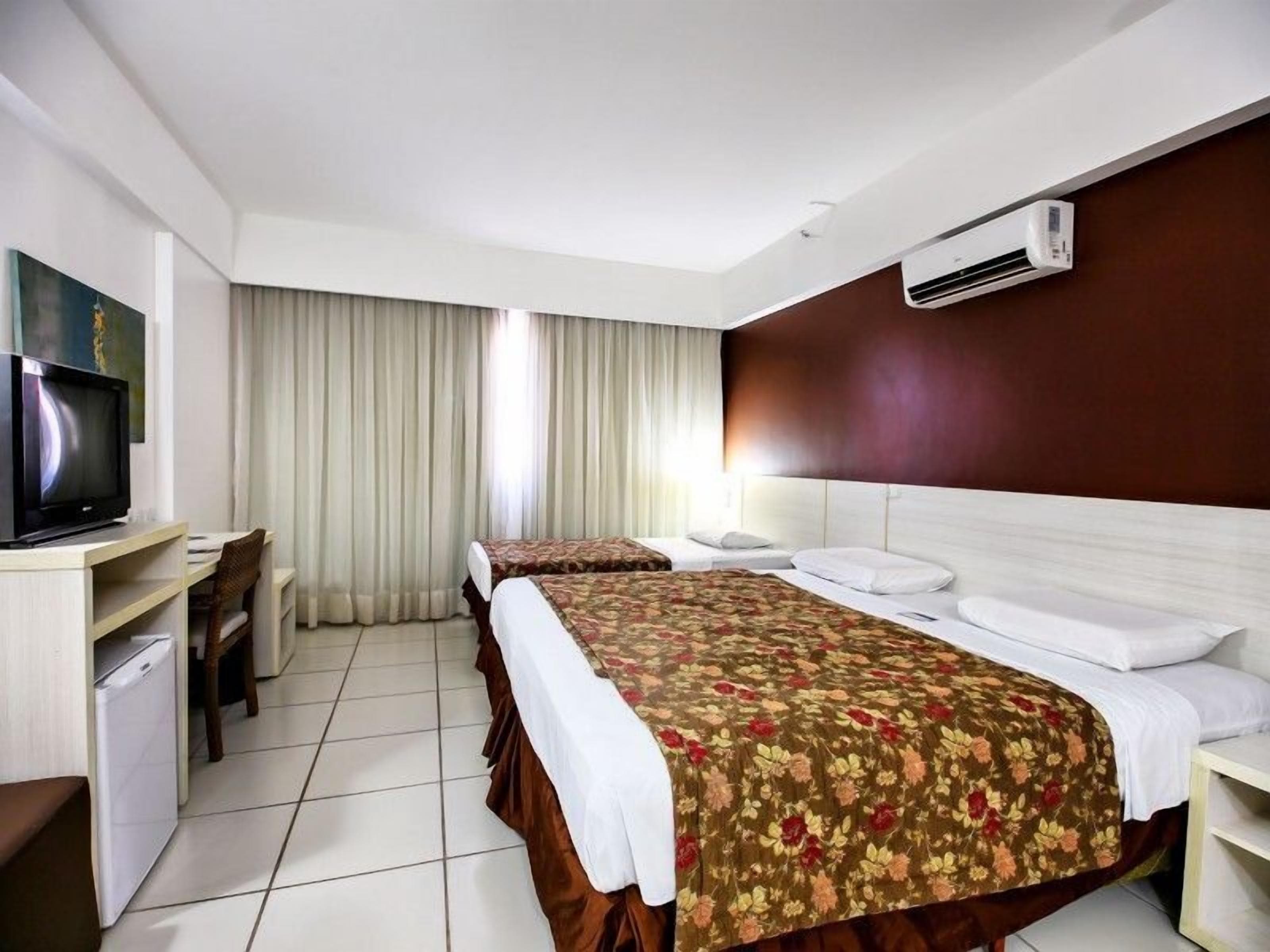 Quarto de hotel com duas camas, uma escrivaninha com televisão e um ar condicionado.