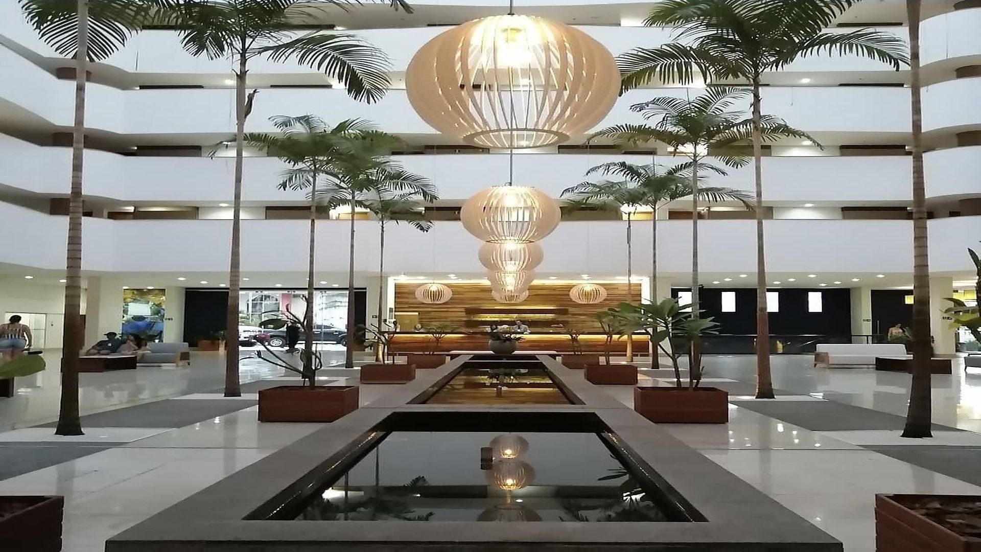 Um hall amplo com uma fonte central, várias palmeiras e um balcão de recepção moderno.