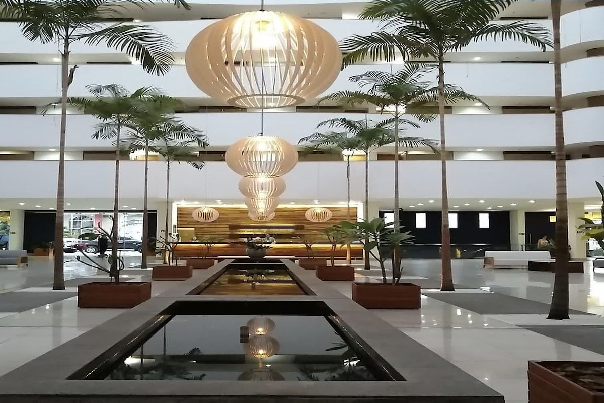 Um hall amplo com uma fonte central, várias palmeiras e um balcão de recepção moderno.