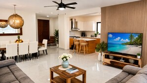 A modern living room with a sofa, a coffee table with a book and flowers, a TV with a tropical beach scene, and a kitchen in the background. - Private Pool Villa 3BR Gated 10 Min Downtown (Punta Cana)
