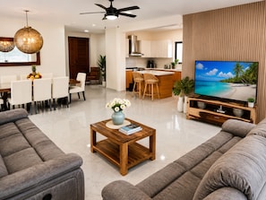 En moderne stue med sofa, sofabord med en bog og blomster, et tv der viser et tropisk strandmotiv, og et køkken i baggrunden.