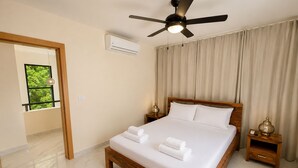 A hotel room with a bed, a ceiling fan, an air conditioning unit, a nightstand, and a window with a view of greenery. - Private Pool Villa 3BR Gated 10 Min Downtown (Punta Cana)