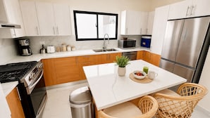 A modern kitchen with white cabinets, stainless steel appliances, a central island, and a dining area with wicker chairs. - Private Pool Villa 3BR Gated 10 Min Downtown (Punta Cana)