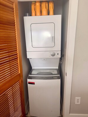 A compact washer and dryer unit in a small utility room with wooden blinds.