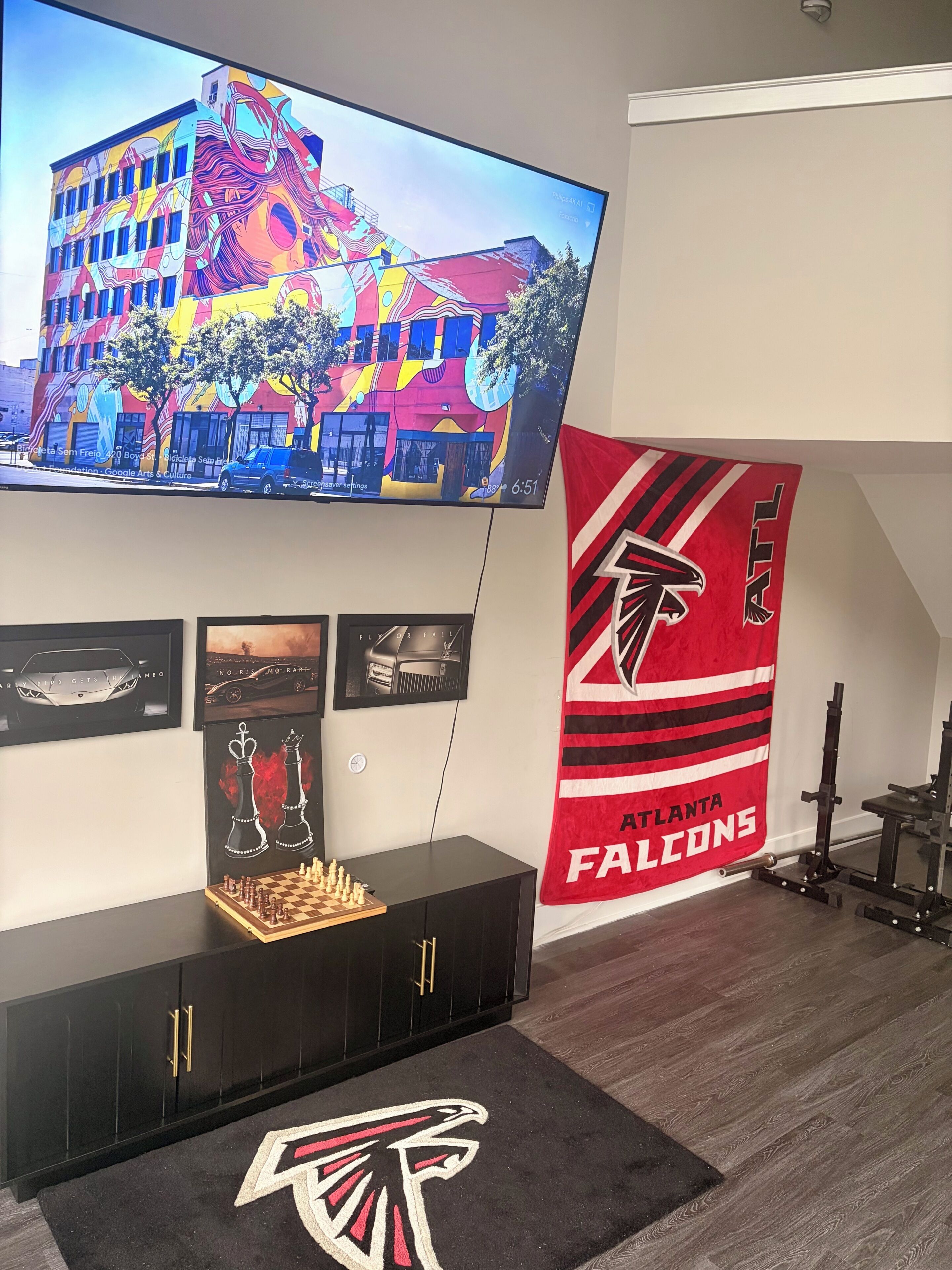 A living room with a television displaying a colorful mural, a chessboard, a gaming console, and a large Atlanta Falcons banner.