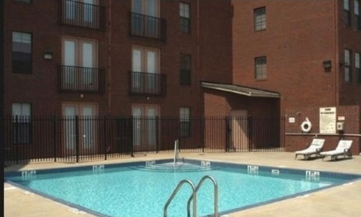 A swimming pool with a metal ladder, surrounded by a fence, and adjacent to a multi-story brick building with balconies.