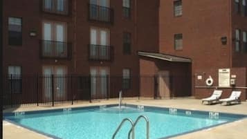 A swimming pool with a metal ladder, surrounded by a fence, and adjacent to a multi-story brick building with balconies.
