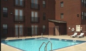 Una piscina con una escalera metálica, rodeada por una cerca y contigua a un edificio de varios pisos con balcones.