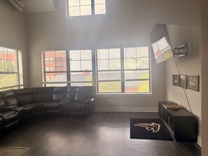 A spacious living room with a sectional sofa, large windows with blinds, a black entertainment unit, and a wall-mounted projector. - Game Day Get Away! Steps From Mercedes Benz stadium! (Atlanta)