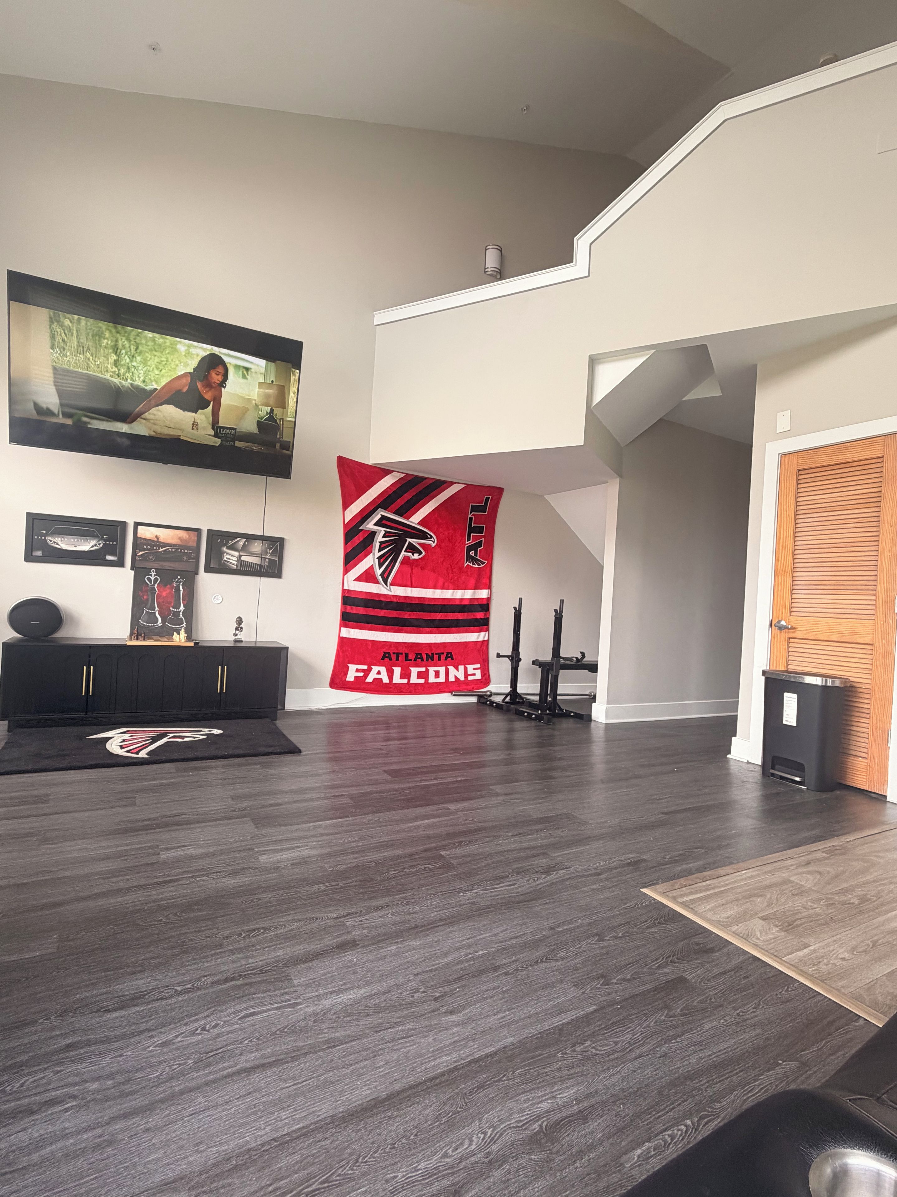 A modern living room with a flat-screen TV mounted on the wall, a black entertainment unit, a red Atlanta Falcons banner, and a wooden door.