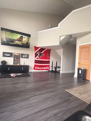 A modern living room with a flat-screen TV mounted on the wall, a black entertainment unit, a red Atlanta Falcons banner, and a wooden door.