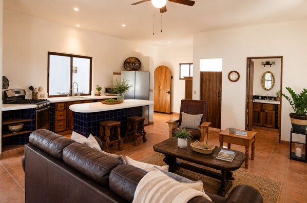 A spacious living room with a brown leather sofa, a wooden coffee table, and a kitchen area with a white countertop and blue tiles.