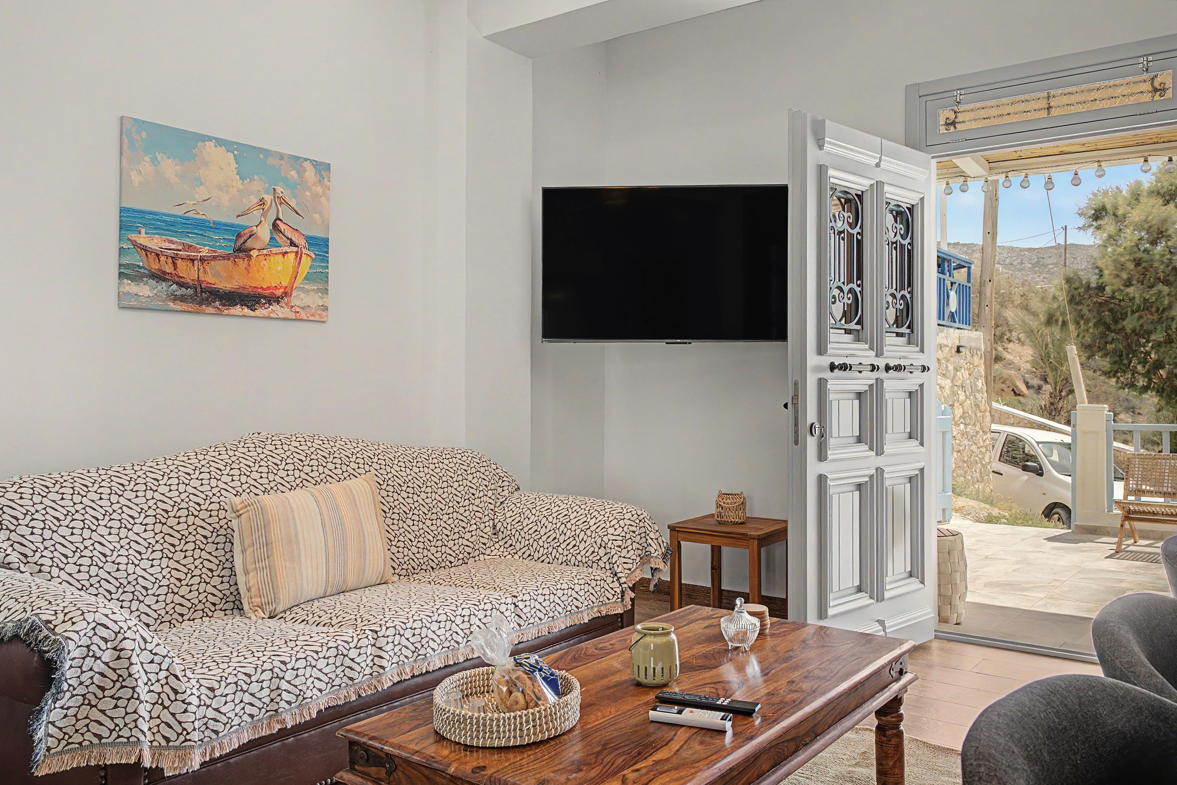 A living room with a patterned sofa, a flat-screen TV, a wooden coffee table with remote controls, and a door leading outside.