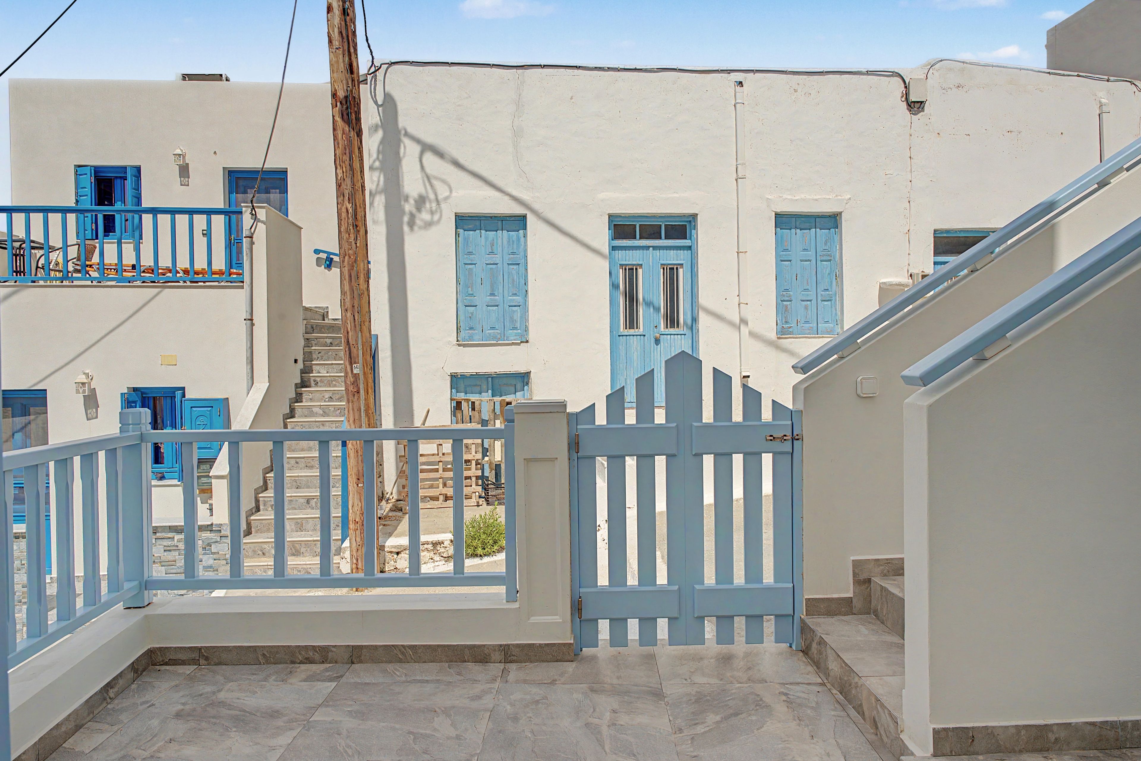 A narrow street with a white building featuring blue doors and windows, a gate, and a staircase leading to an upper level.