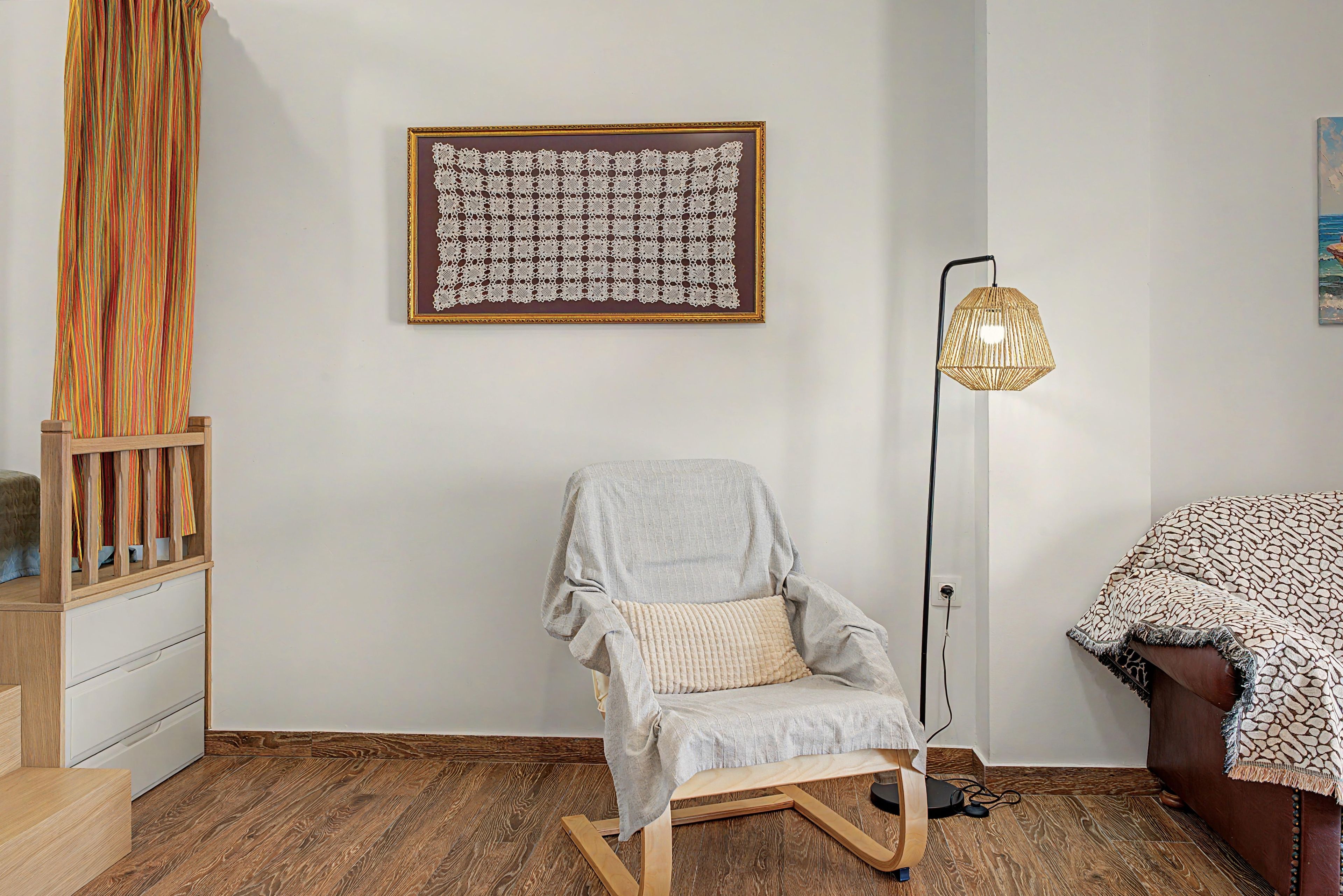 Une pièce avec un plancher en bois, une chaise avec un coussin, un lampadaire et une œuvre d’art encadrée accrochée au mur.
