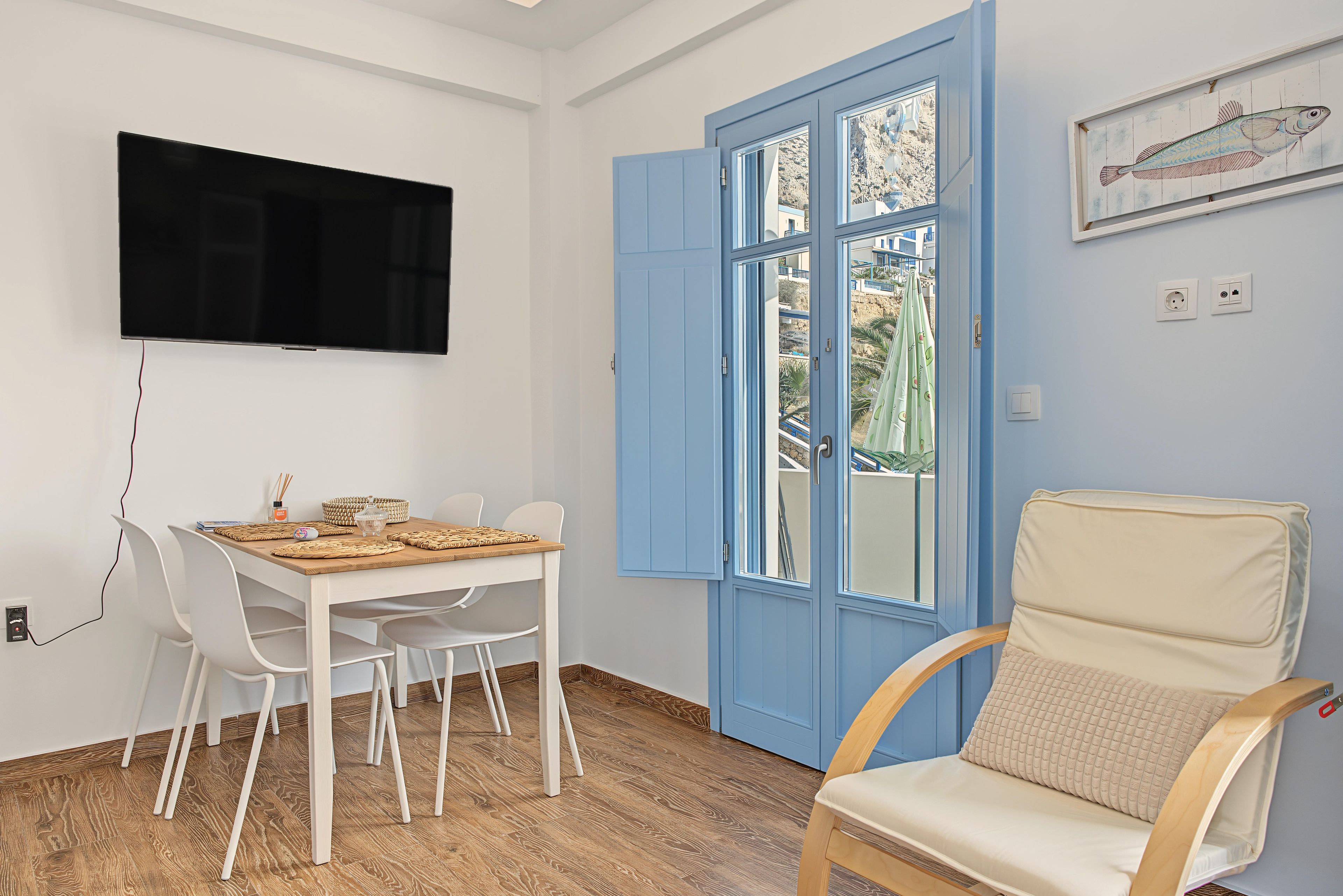 Ein Zimmer mit an der Wand befestigtem Fernseher, einem Esstisch mit Stühlen, einer blauen T ür, einem Bett mit Kissen und einer Wandsteckdose.