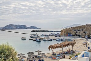 A coastal marina with boats, a sandy beach with umbrellas, and a rocky hillside in the background.