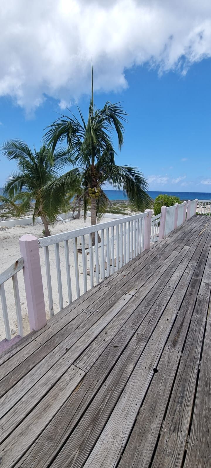 Une promenade en bois, agrémentée de rampes de garde blanches, qui mène à une plage de sable fin, bordée de palmiers, avec l’océan en toile de fond.