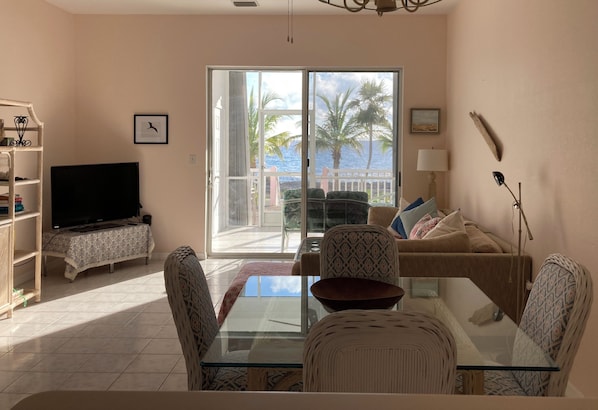 A living room with a glass table, sofa, and chairs, a television, and a view of palm trees through a sliding glass door.