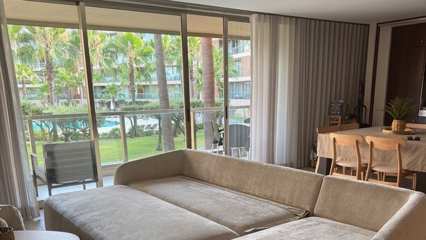 Un salon moderne avec un canapé d’angle, une petite table ronde et une vue sur une piscine et la verdure environnante.