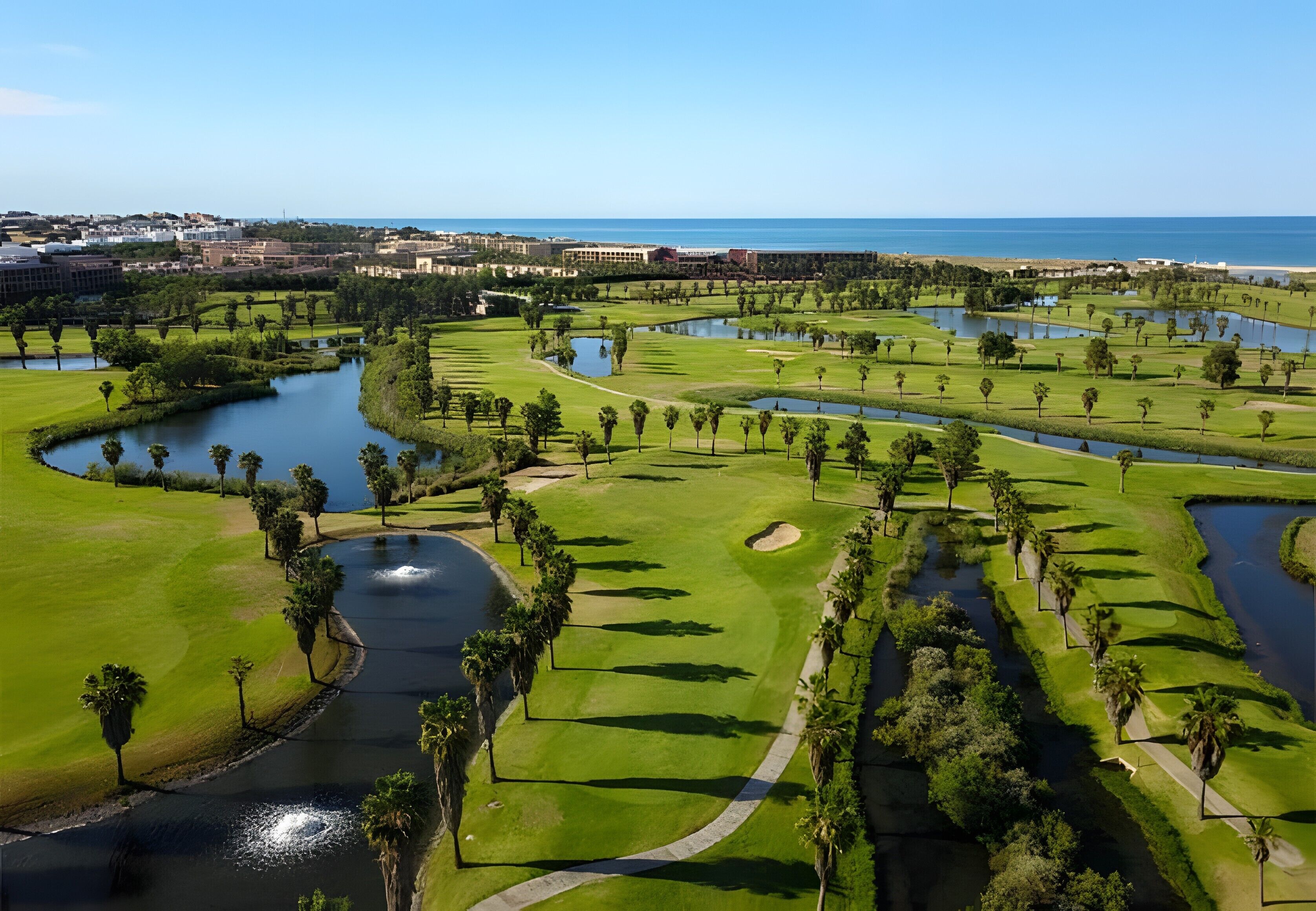 Ein Golfplatz mit mehreren Wasserhindernissen, umgeben von Grünflächen und Palmen, mit Blick auf den Ozean im Hintergrund.